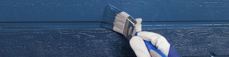 Käsineellinen käsi siveltimellä maalaa puupaneelin ulkoseinää tummansiniseksi.