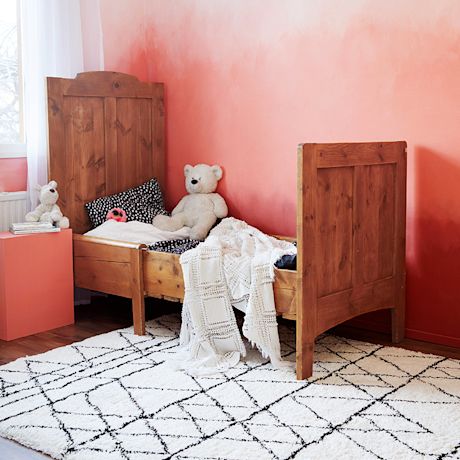 Roosaseinän edessä puuvuode, pehmolelut, valkoinen virkattu peitto ja mustavalkoinen matto