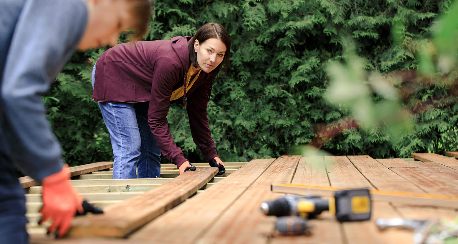 Kaksi ihmistä asettaa puisia terassilautoja vihreää pensasaitaa vasten.