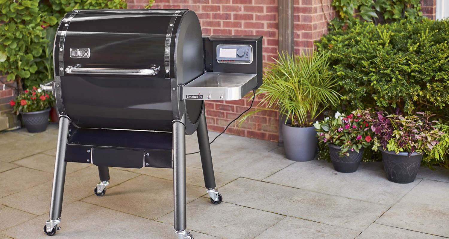 Musta Weber SmokeFire -grilli pihalaatoilla tiiliseinää vasten, vieressä kukka- ja ruukkuistutuksia.