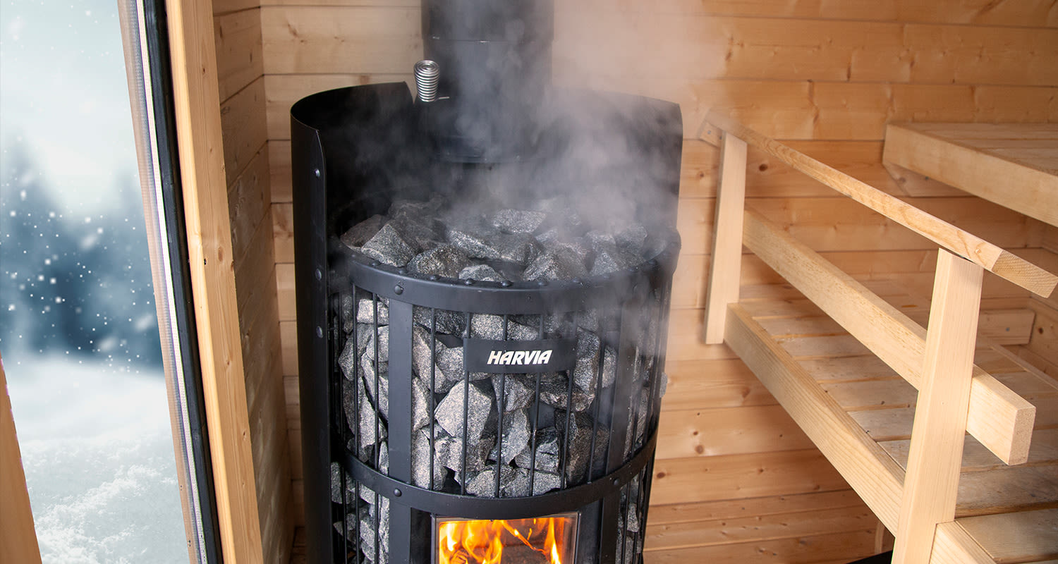 Mustan Harvia-kiukaan höyryävät kivet puusaunassa, lauteet oikealla ja luminen metsä ovesta.