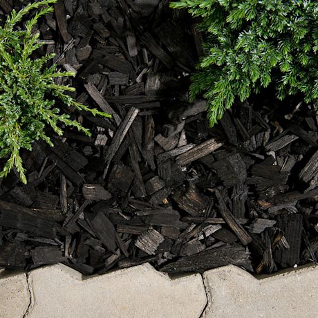 Musta puuhake kattaa pinnan, yläreunassa vihreitä havunoksia ja alhaalla vaalea kiveys