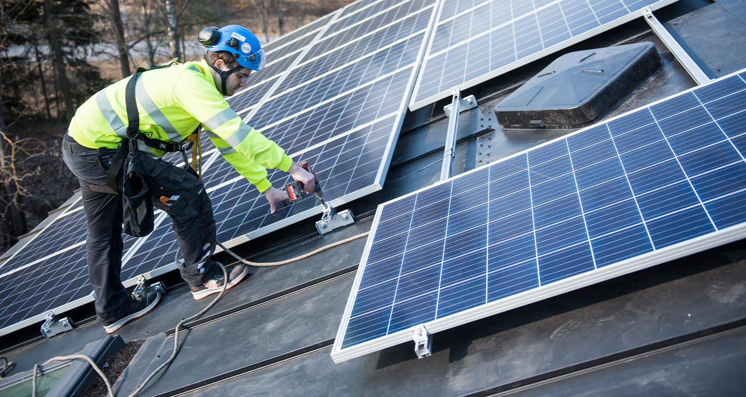 Työntekijä keltaisen huomiotakin ja sinisen kypärän kanssa asentaa aurinkopaneeleja katolle