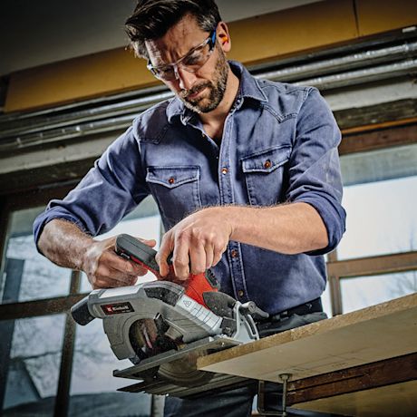 Mies sinisessä farkkupaidassa sahaamassa puulevyä punaisella käsisahalla valoisassa verstaassa