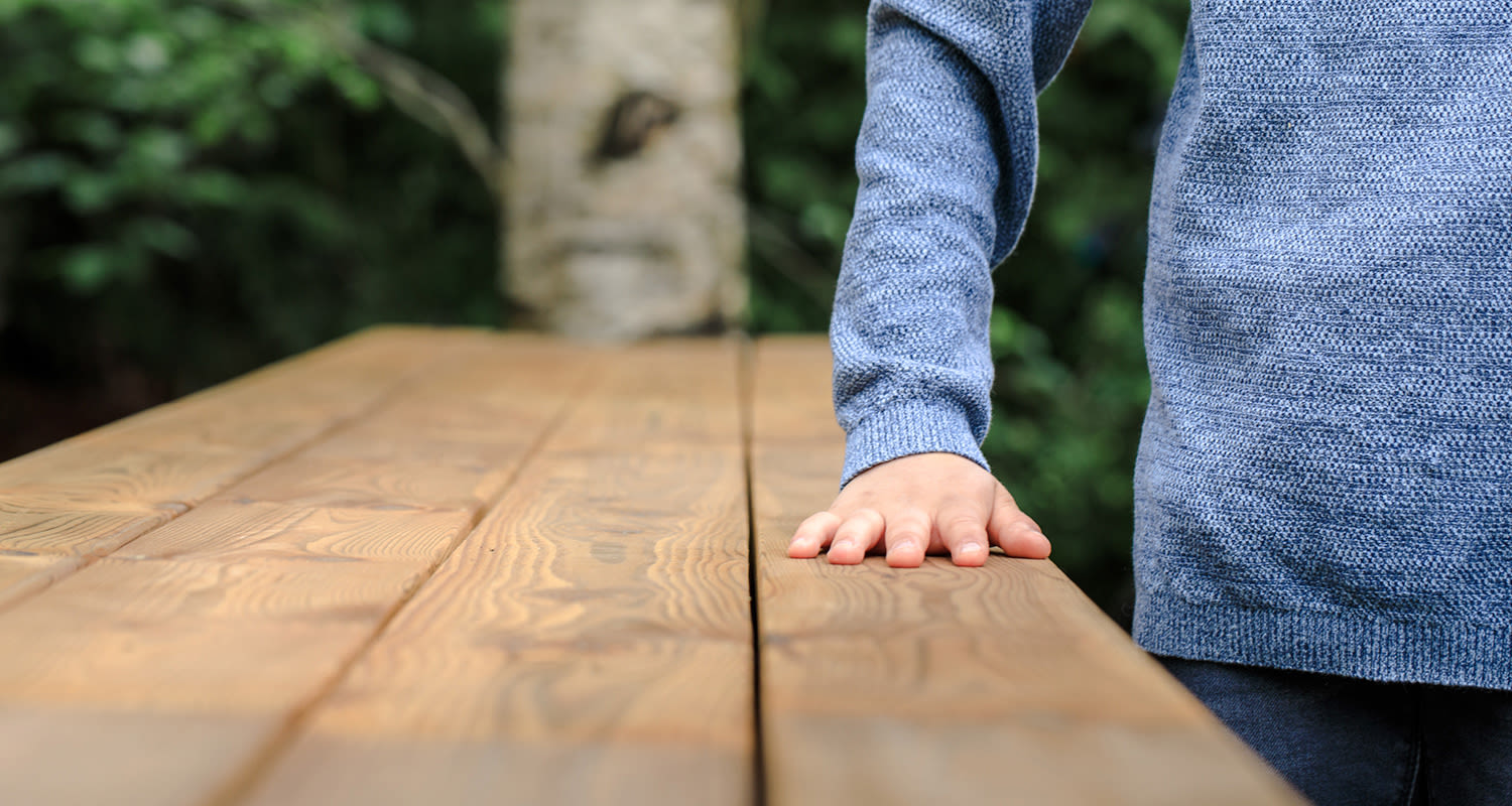Siniseen neuleeseen pukeutuneen henkilön käsi nojaa puupöytään metsätaustalla