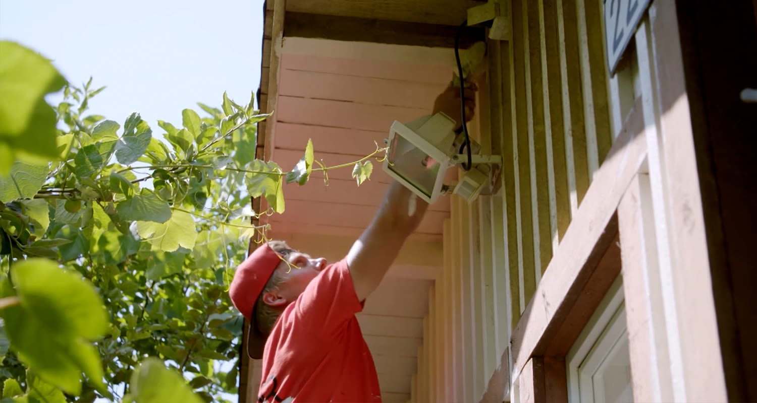 Punapaitainen mies asentaa valkoista ulkovaloa vaaleanpunaisen räystään alle.