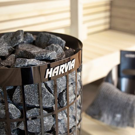 Musta Harvia-kiuas kivikorissa täynnä harmaita kiviä vaalean puusaunan lauteiden edessä