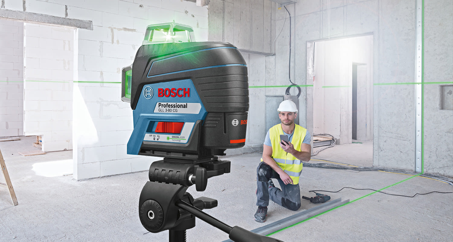 Bosch-laseri jalustalla vihreine linjoineen keskeneräisessä sisätilassa, työntekijä kännykällä