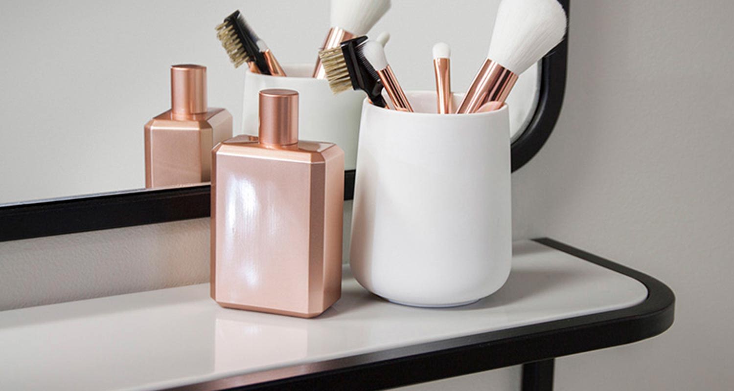 Peilin edessä valkoinen meikkiteline, jossa ruusukultaisia siveltimiä ja ruusukultainen pullo