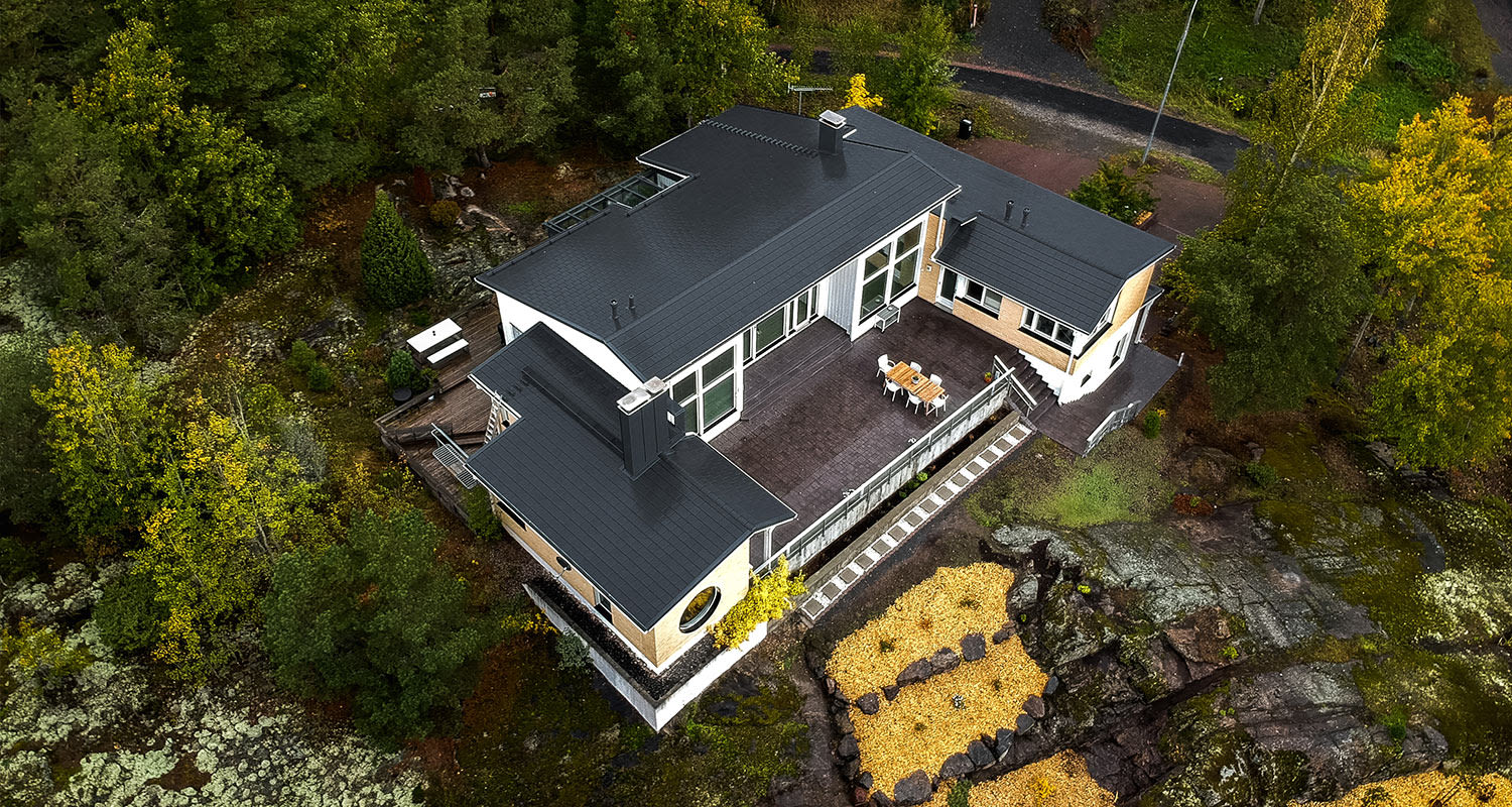 Ilmakuva modernista talosta harmaalla katolla ja terassilla keltavihreän syksymetsän keskellä.