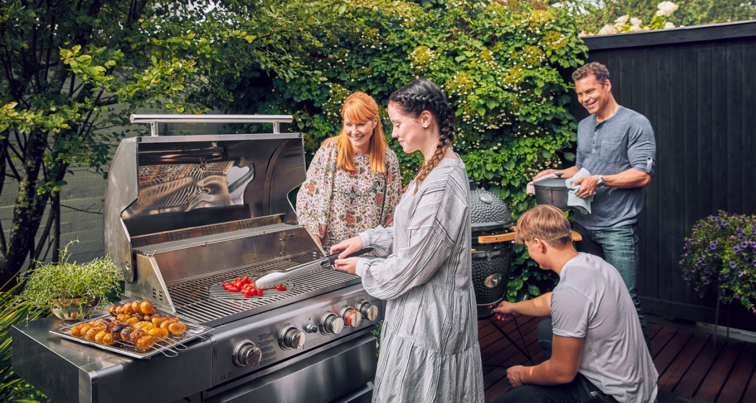 Neljä ihmistä grillaa metalligrillillä puutarhaterassilla, vartaita ja punaisia paprikoita.