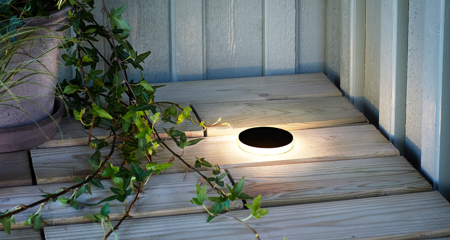 Pyöreä mustakantinen LED-valaisin loistaa puulaudoitetulla terassilla köynnöksen vieressä.