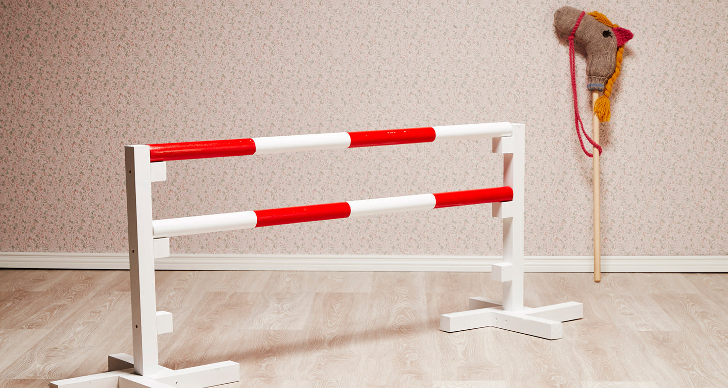 Valko-punainen ratsastuseste puulattialla kukkaseinää vasten, seinässä virkattu keppihevonen