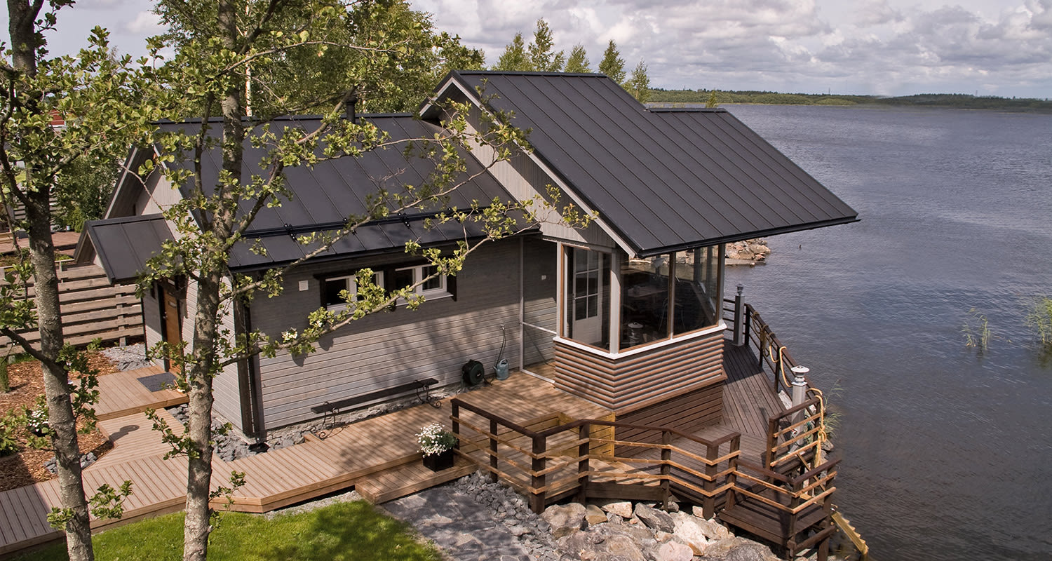 Harmaalankainen mökki mustalla peltikatolla puisella terassilla järven rannalla kesämaisemassa