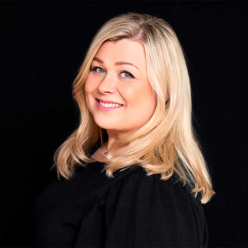 Muotokuva vaaleahiuksisesta henkilöstä kaulakorussa ja mustassa paidassa mustalla taustalla