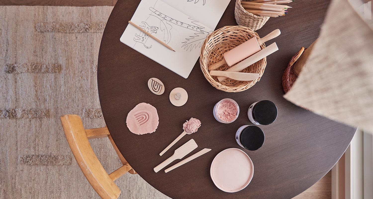 Ylhäältä: tumma pyöreä pöytä, korissa puiset työkalut, vaaleanpunainen muovailuvaha ja maalipurkki