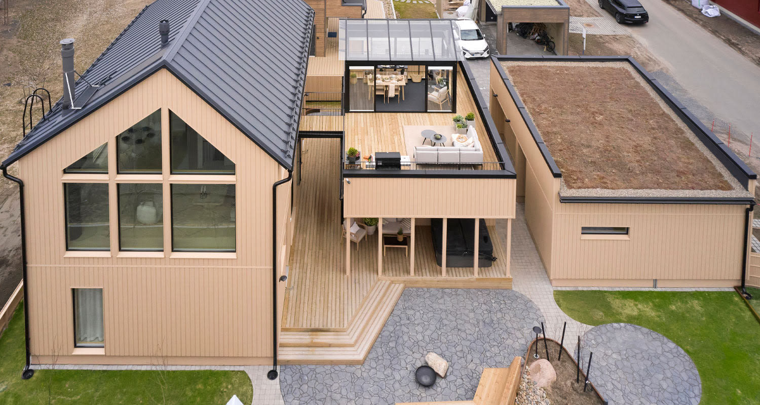 Ilmakuva beigest puisesta talosta mustakattoisesta, lasikatettu terassi sohvaryhmällä ja nurmikenttä