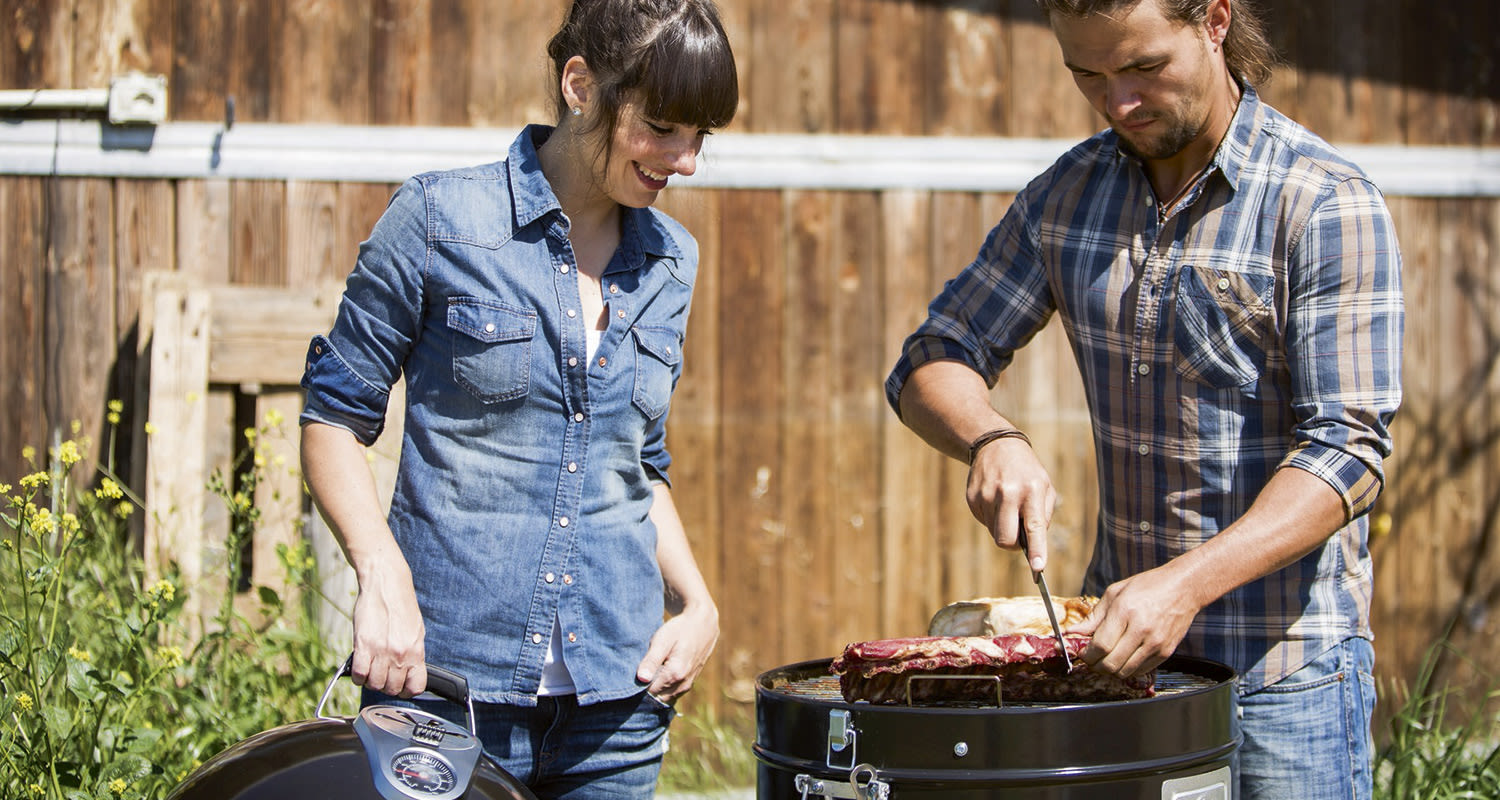 Mies leikkaa ribsejä savustimella ja nainen pitelee sen kantta pihalla.