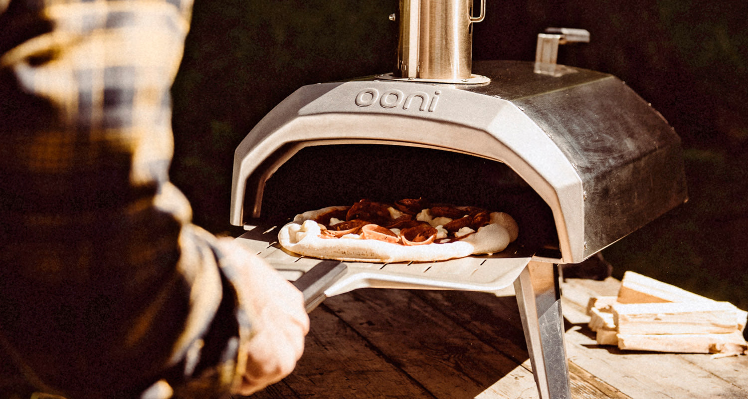 Ulkona hopeinen Ooni-pizzauuni, käsi työntää pepperonapizzaa sisään, halkopino vieressä.