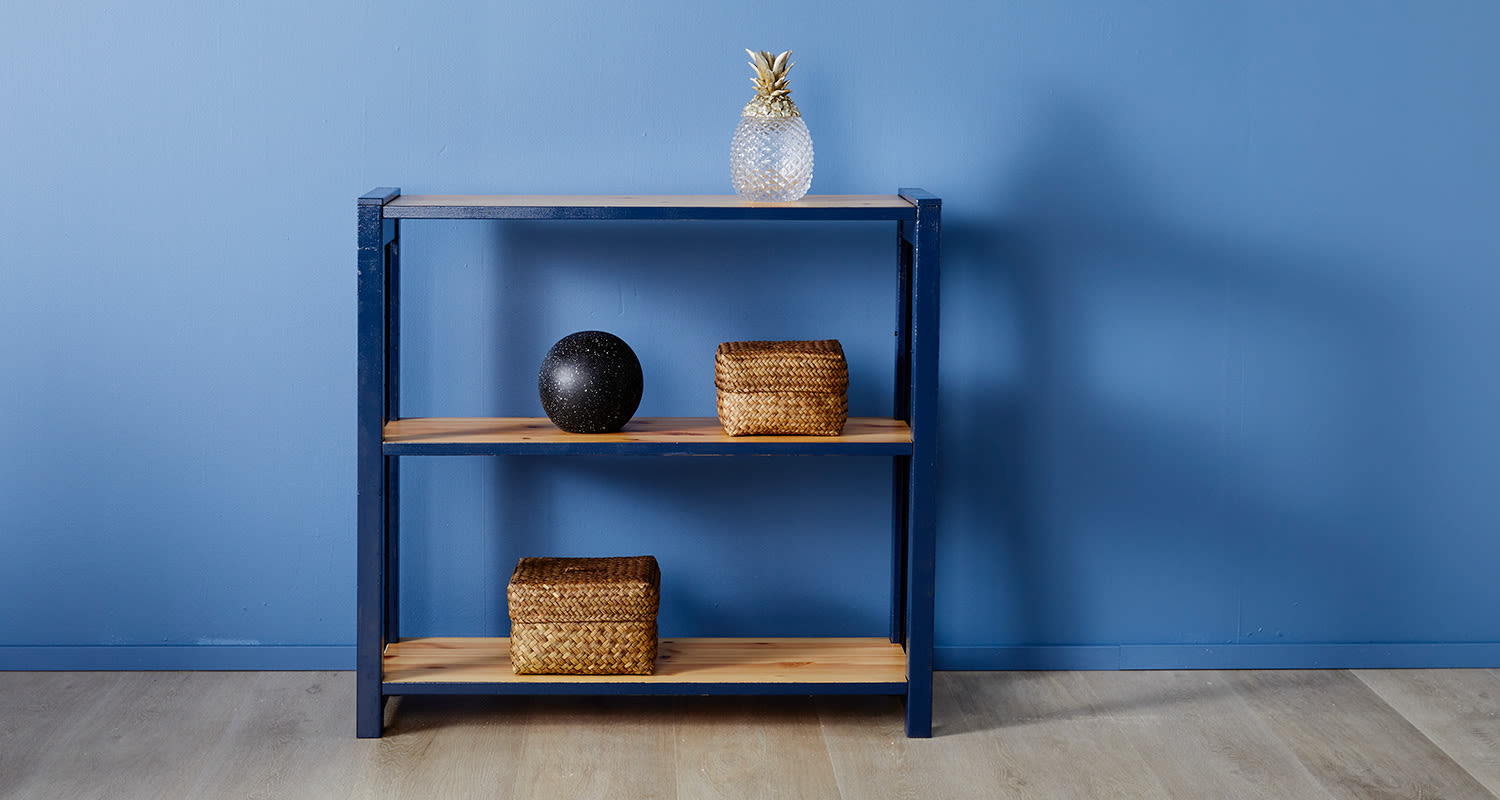 Sininen kolmitasoinen hylly sinisellä seinällä: lasinen ananasilamppu, musta pallo ja punoskorit