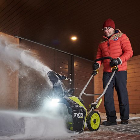 Henkilö käyttää Ryobi RY18STX45A-240 18V ONE+ -lumilinkoa rakennuksen edustalla, ja laite puhaltaa lunta tehokkaasti sivulle. Lumilingossa on kirkas työvalo, joka valaisee työskentelyaluetta.
