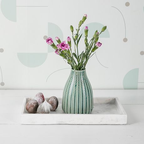 Vihreä kuvioitu maljakko vaaleanpunaisine neilikoin, tarjottimella kiviä ja höyhen.