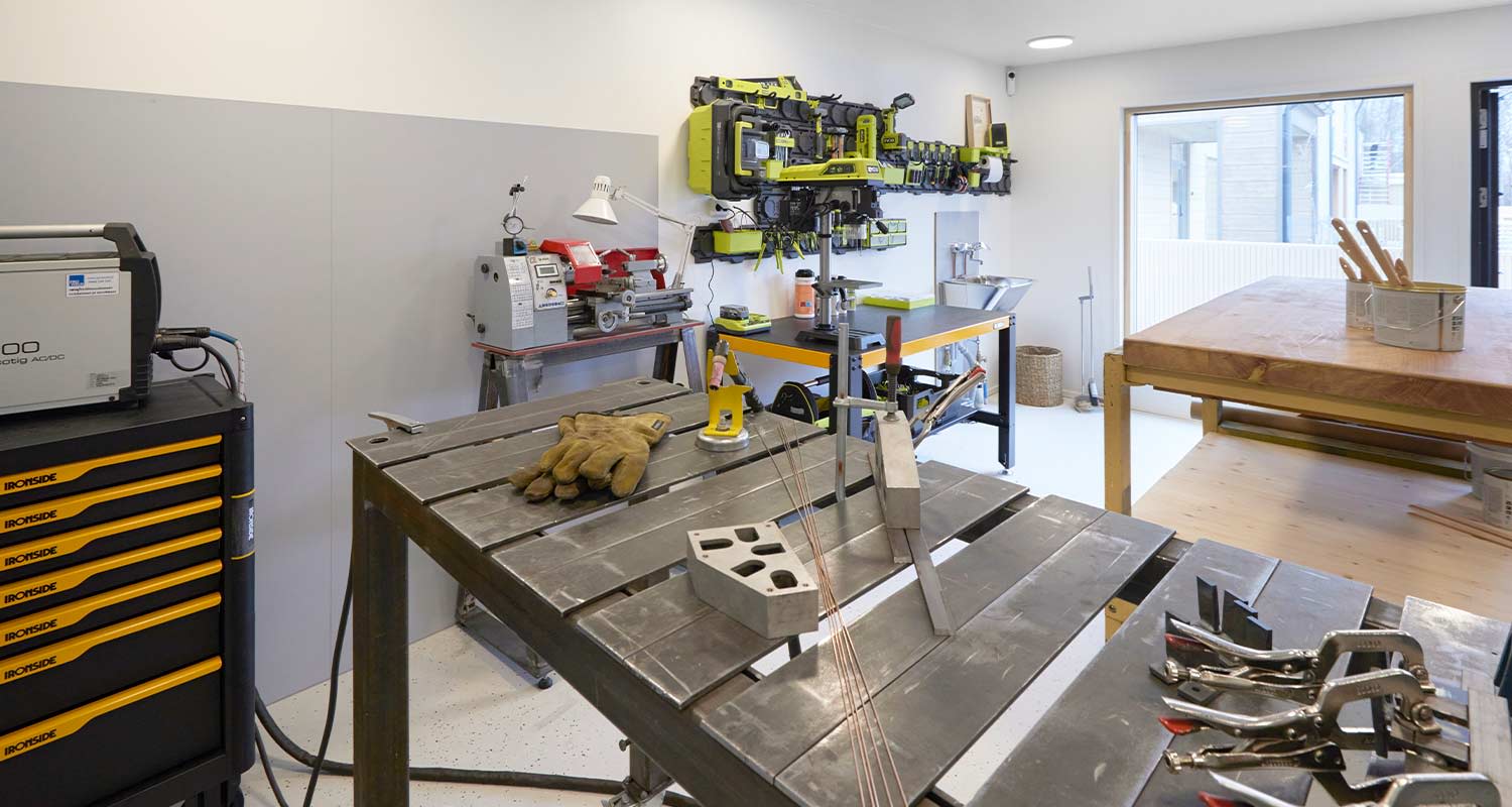 Valoisassa työhuoneessa metallipöytiä, keltamustia työkaluja, suojakäsineet ja ikkunanäkymä.