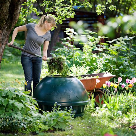 Nainen työntää ruoho- ja kasvimassaa vihreään maapallokompostoriin vehreässä puutarhassa.