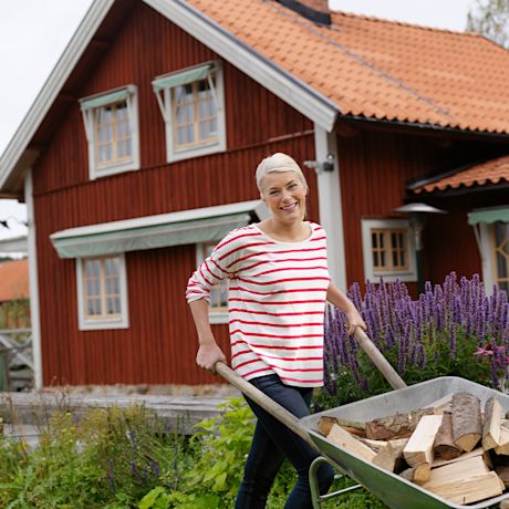 Nainen työntää puukuormaa kottikärryillä mökin pihalla.