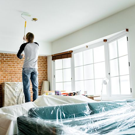 Mies maalaa valkoista kattoa olohuoneessa, tiiliseinä ja muovipeitteinen sohva näkyvissä.