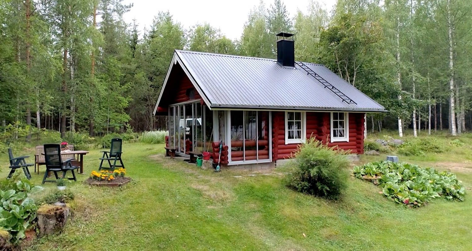 Punainen pihamökki harmaalla katolla metsän laidassa, nurmella tuolit, pöytä ja kukkapenkki