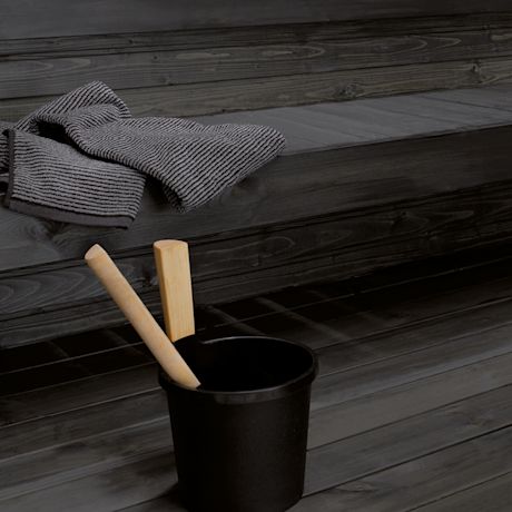 Harmaa raitapyyhe tumman puupenkin päällä, musta sauna-ämpäri ja vaaleat puukauhat puulattialla.