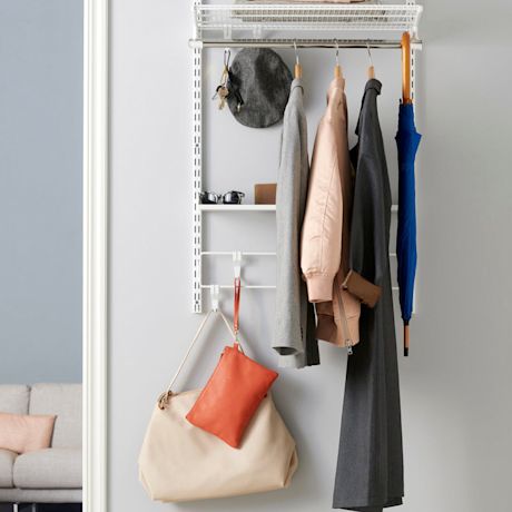 Seinänaulakossa harmaa ja beige takki, oranssi pussukka, sininen sateenvarjo vaaleaa taustaa vasten