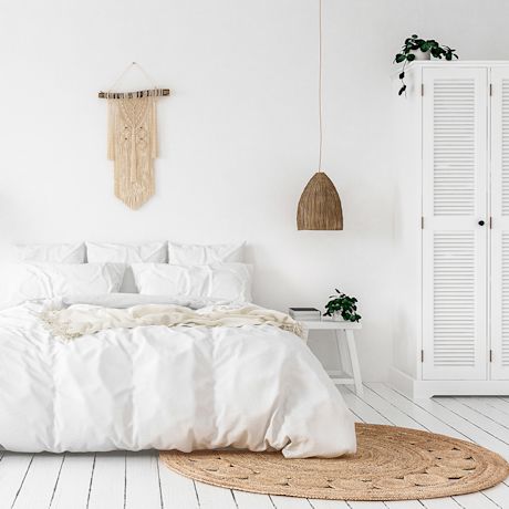 Valkoinen minimalistinen makuuhuone: sänky juuttimatolla, rottinkivalaisin ja makrameekoriste.