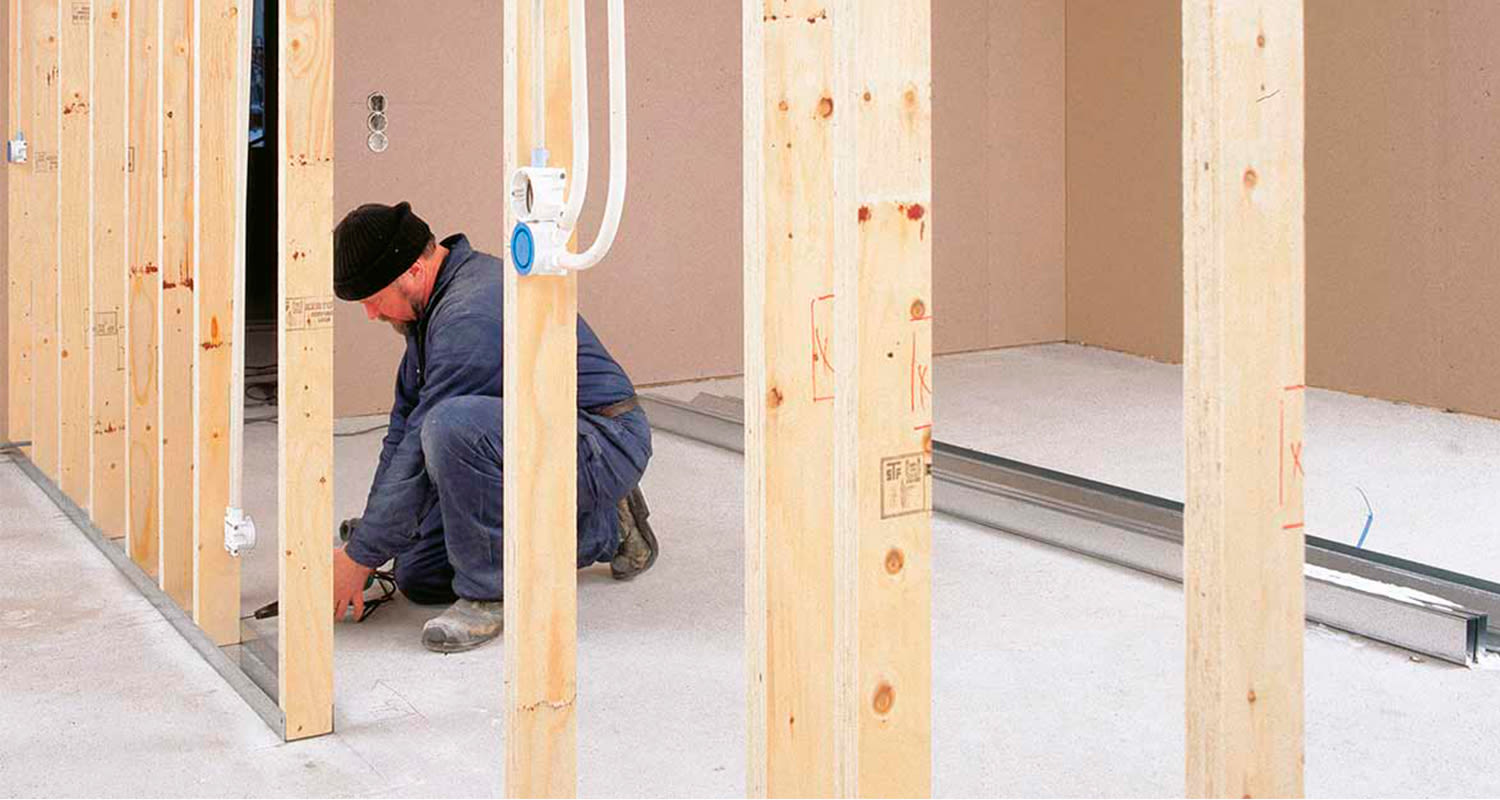 Työmies asentaa vaaleista puutolpista sisäseinäkehikkoa harmaalle betonilattialle.