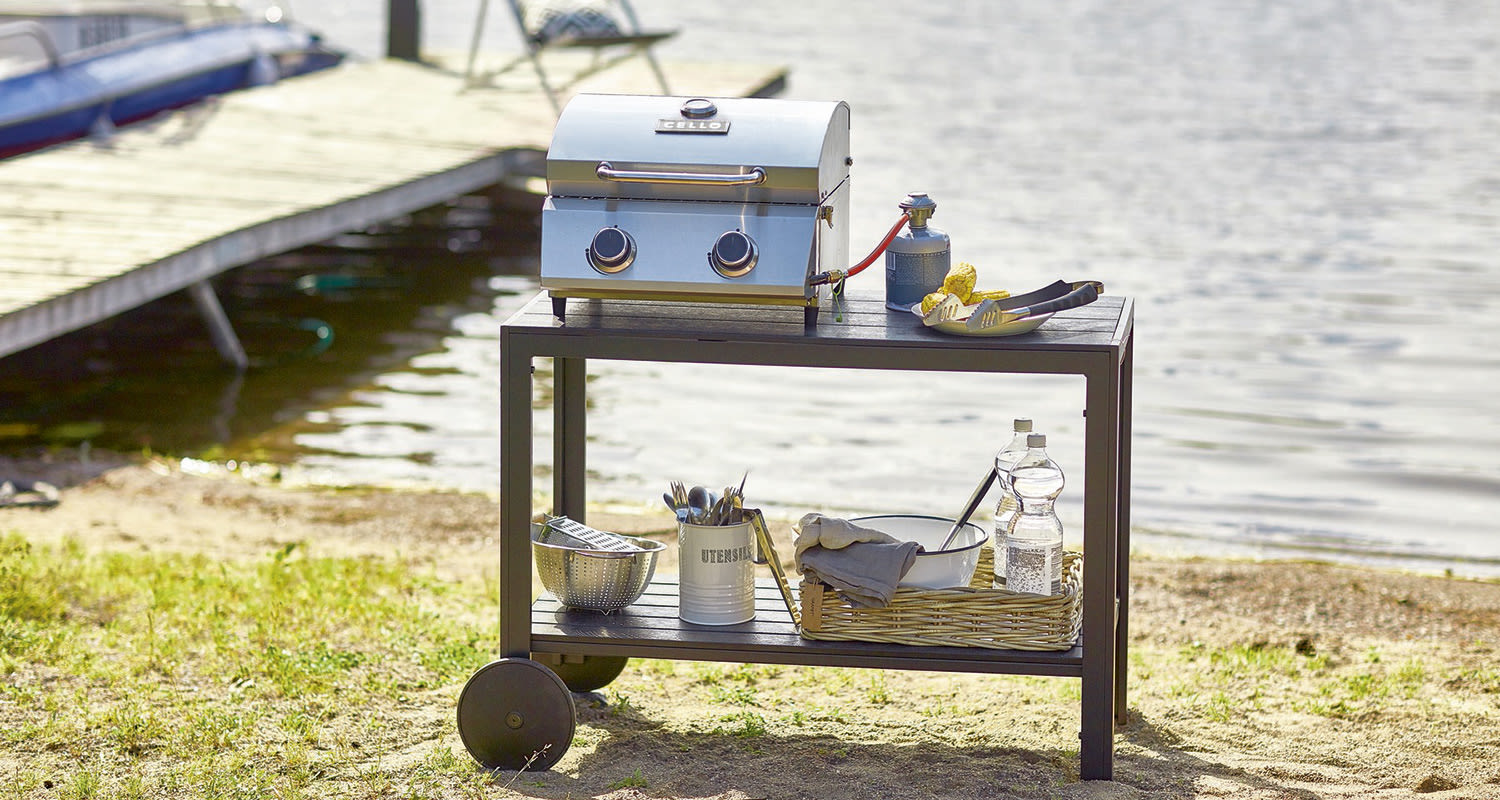 Musta rullallinen alusta laiturin rannalla, päällä hopeinen kaasugrilli ja ruokatarvikkeita.