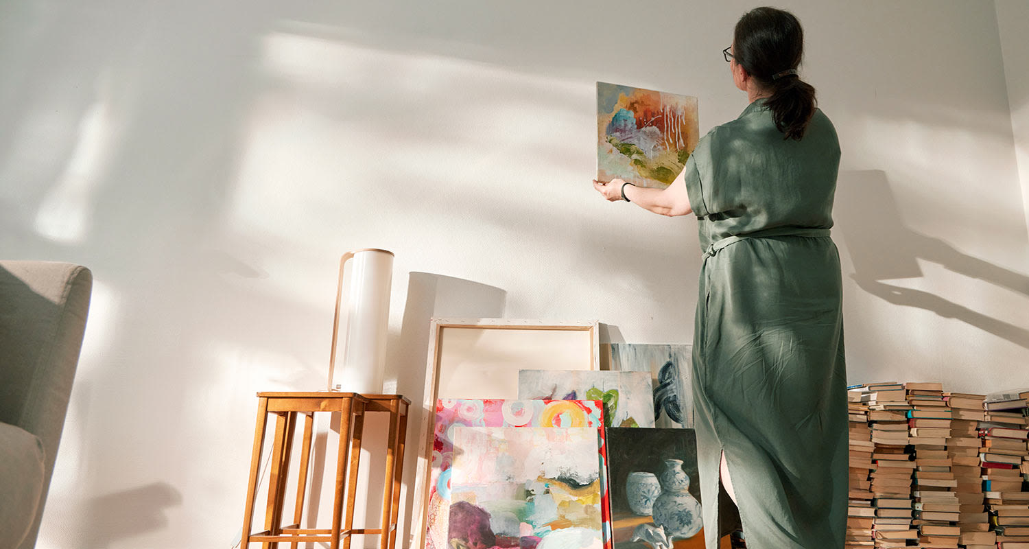 Nainen vihreässä mekossa ripustaa abstraktitaulun valkoiselle seinälle, edessä taulu- ja kirjakasa.