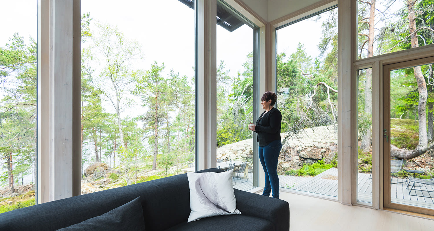 Vaalea puinen mökki lattiasta kattoikkunoin tumma sohva valkoisella tyynyllä metsämaiseman äärellä