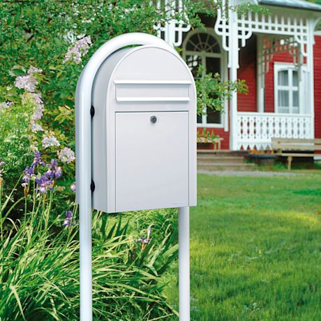 Valkoinen postilaatikko jalustassa nurmikolla, taustalla punavalkoinen puutalo ja kurjenmiekkoja