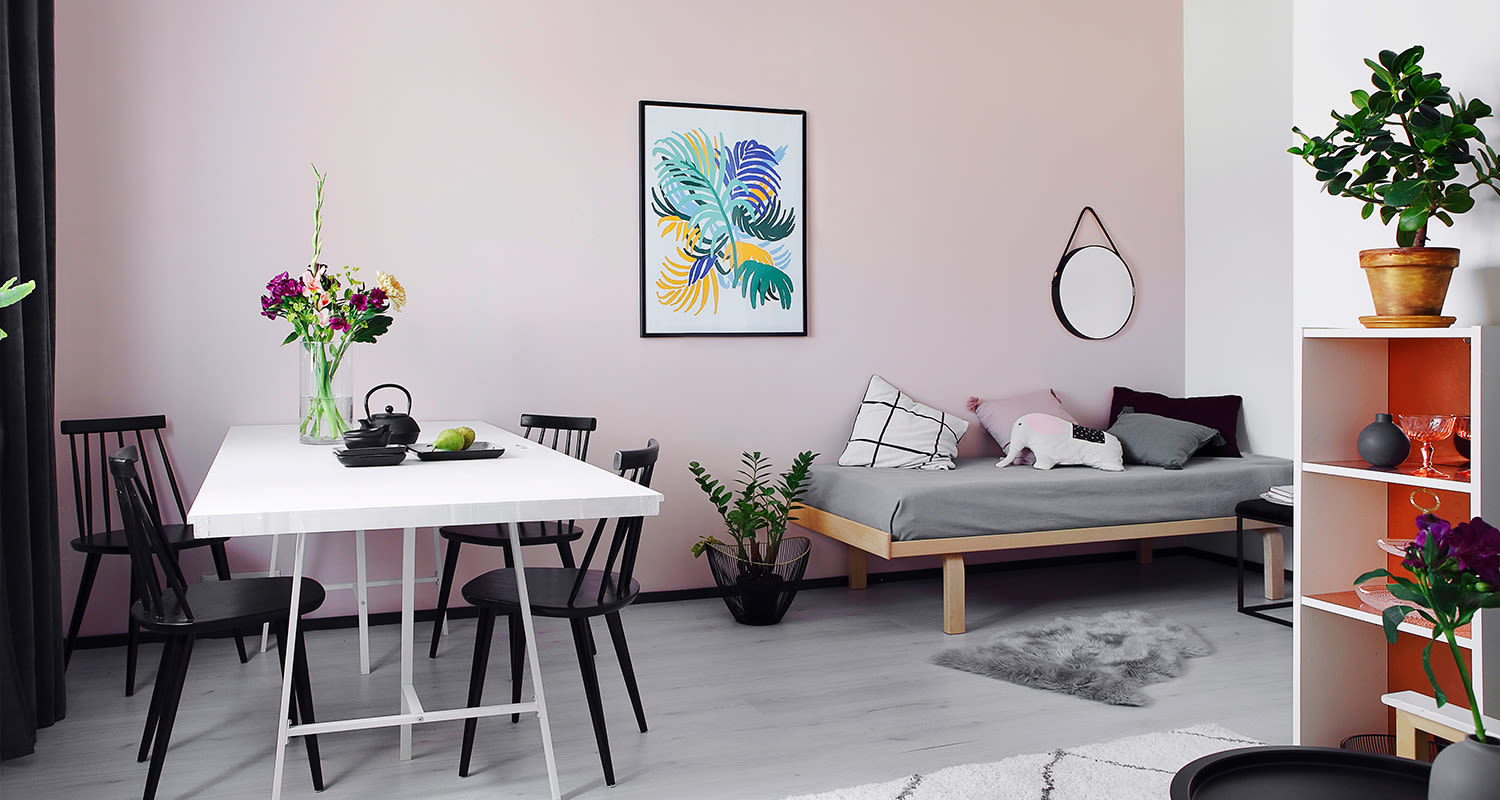 Vaaleanpunainen huone: valkoinen pöytä mustilla tuoleilla, kukkakimppu ja harmaa vuodesohva.