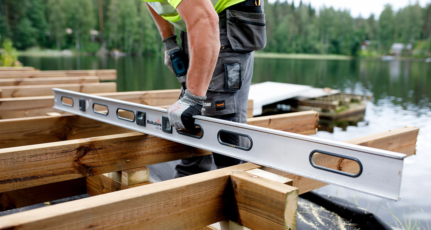 Rakentaja neonkeltaisessa paidassa tarkistaa vesivaakalla puupalkkien tasoa järven rannalla