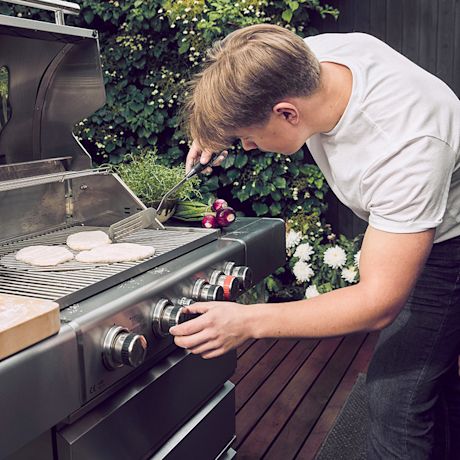 Henkilö kääntää puuterassilla teräksisen kaasugrillin ritilällä paistuvia pihvejä.