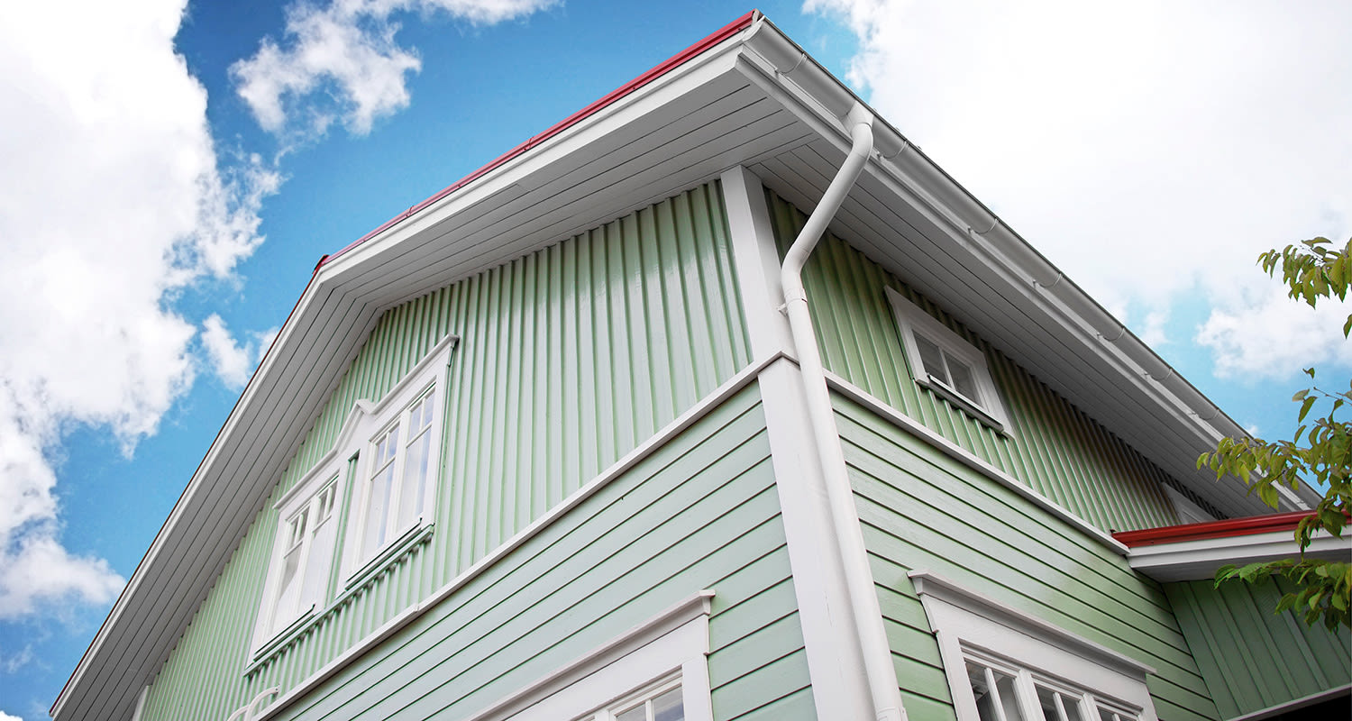 Vihreän puutalon kulma, valkoiset ikkunat ja räystäät, punainen katto, sinitaivas.