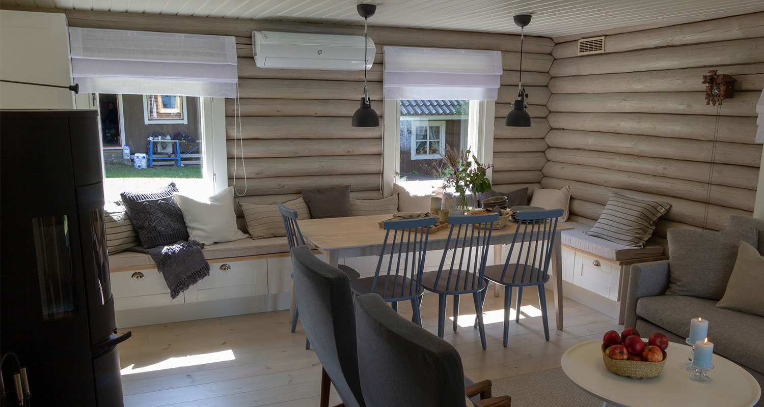 Vaaleassa hirsimökissä puinen ruokapöytä sinisine tuoleineen ja ikkunapenkit tyynyillä