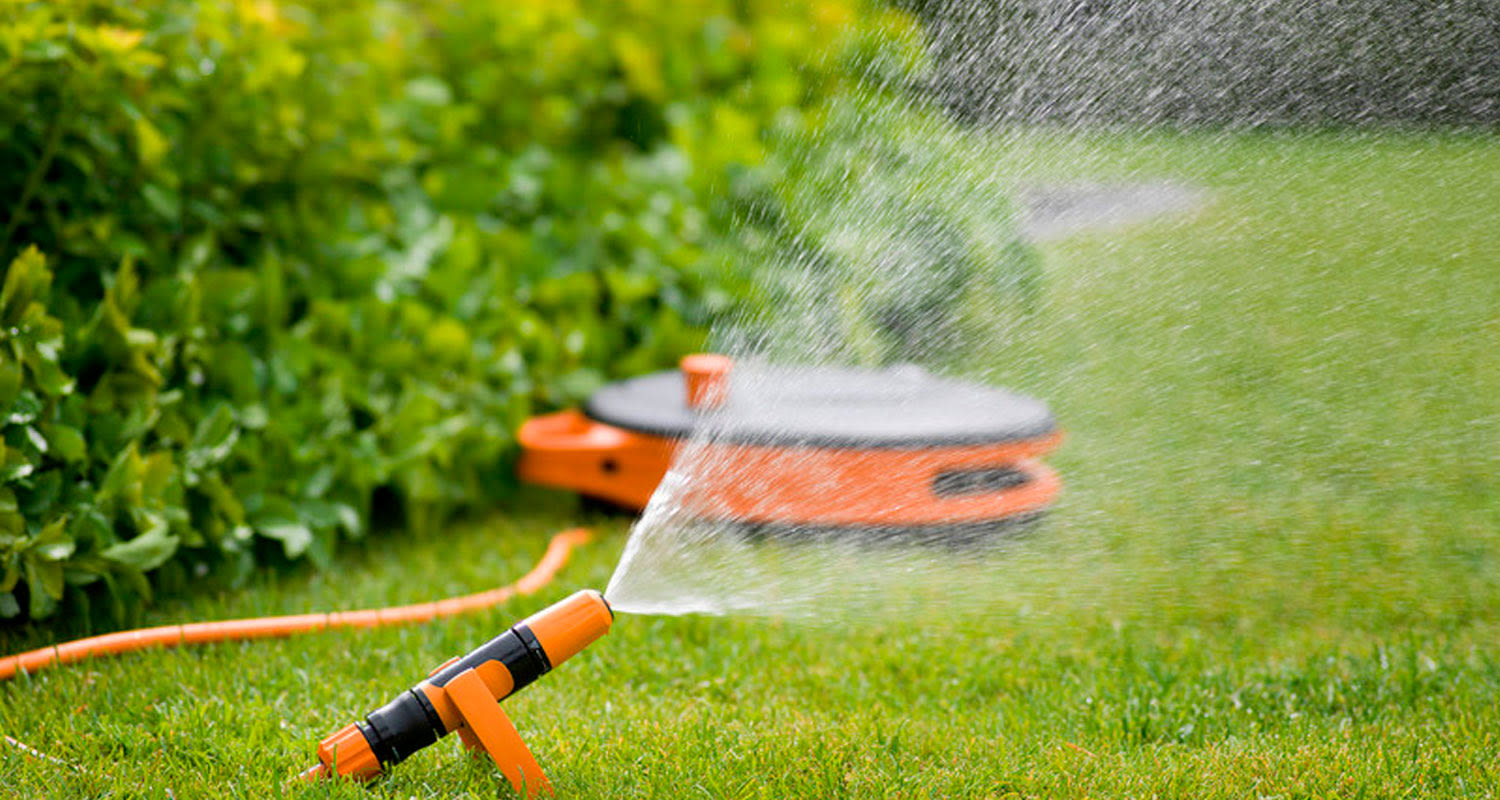 Oranssi-musta pyöriväsuihku suihkuttaa vettä vihreälle nurmikolle pensasaidan edessä.