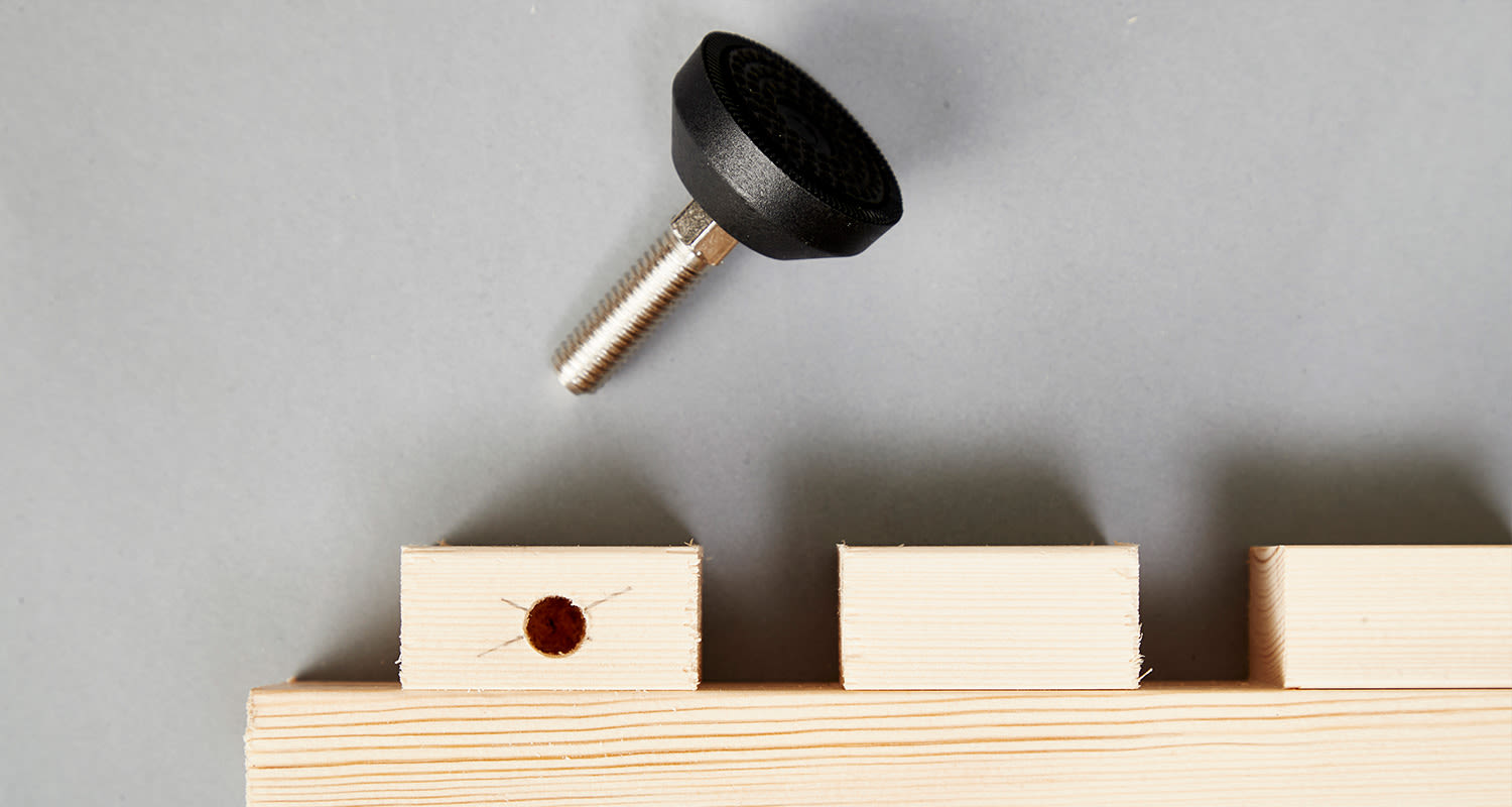 Harmaa tausta, metallinen säätöruuvi mustalla kahvalla ja kolme puupalikkaa, joista yksi rei’itetty