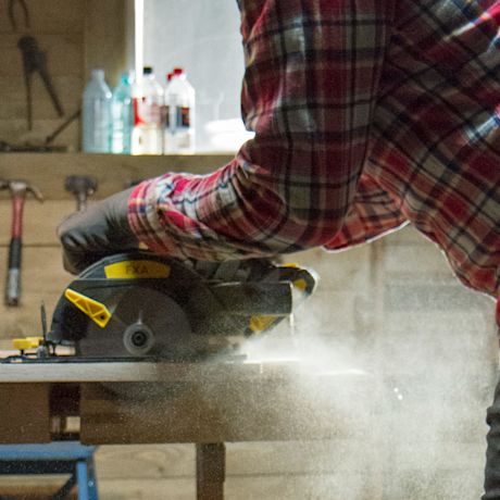 Ruudulliseen paitaan pukeutunut henkilö sahaa puuta pöytäsahalla työpajassa, puupöly leijuu.