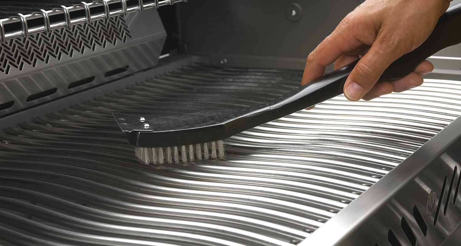 Käsi puhdistaa mustankahvaisella teräsharjalla aaltoilevaa ruostumatonta grilliritilää