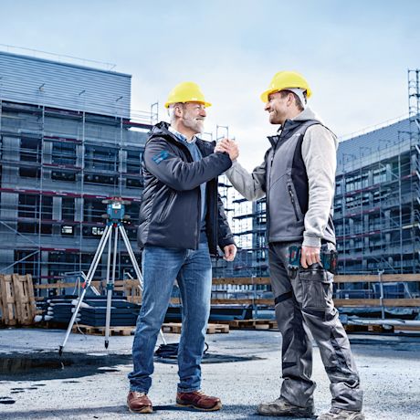 Kaksi rakennusmiestä kättelee keltaisissa kypärissä työmaalla, taustalla telineet.