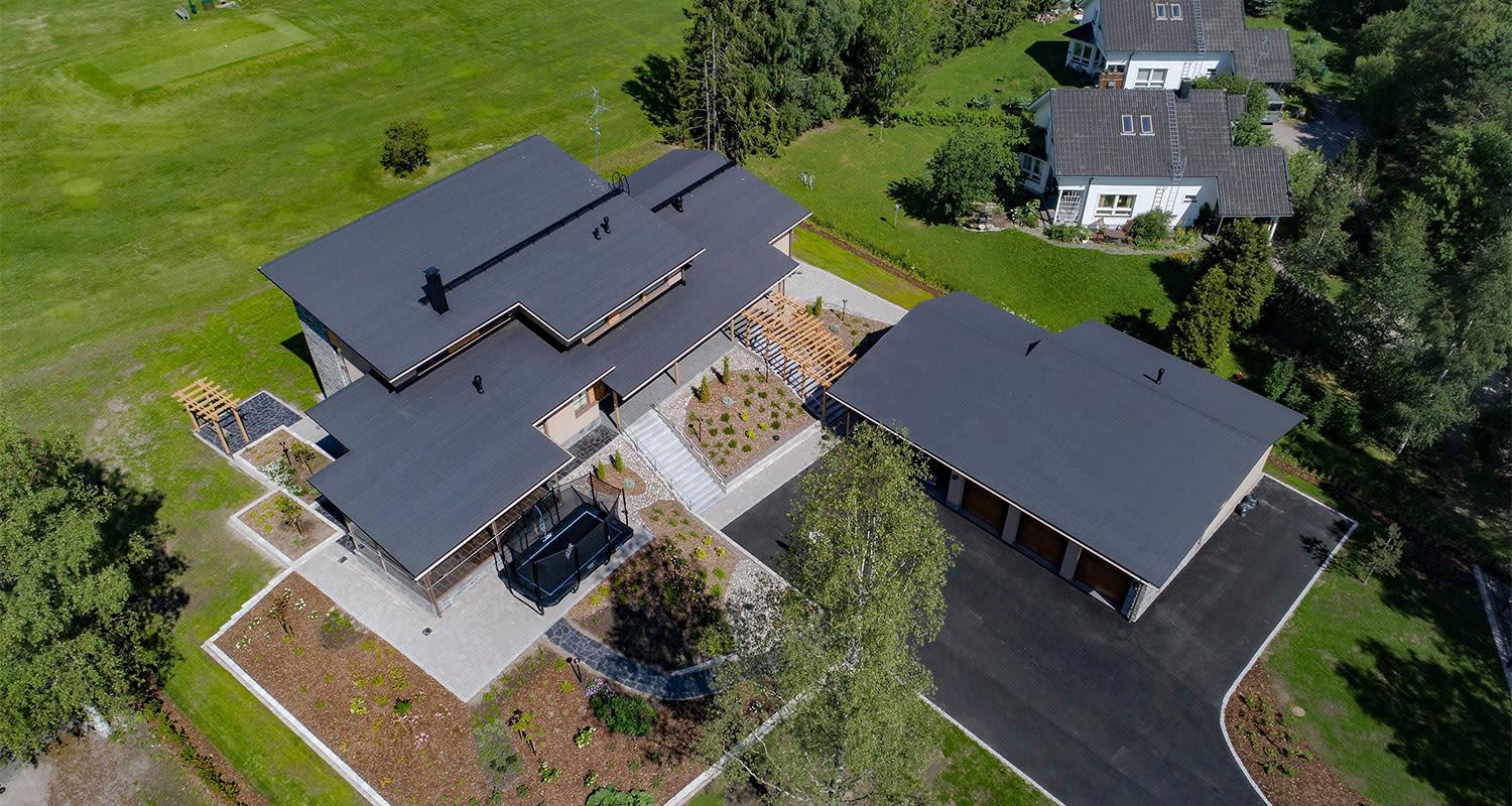Ilmakuva modernista mustakattoisesta talosta ja erillisestä autotallista laajoilla nurmipihalla